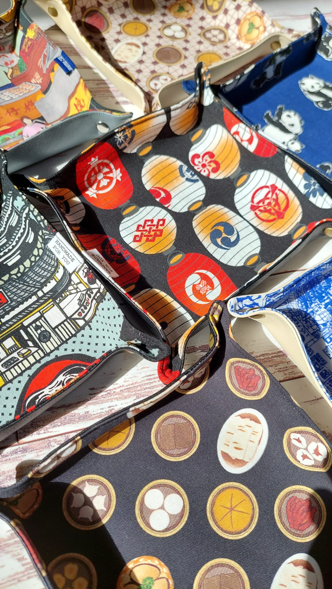 A collection of handmade square catchall trays featuring various fabric patterns, including traditional Japanese lanterns, panda, dim sums, all finished with snap-button and faux leather