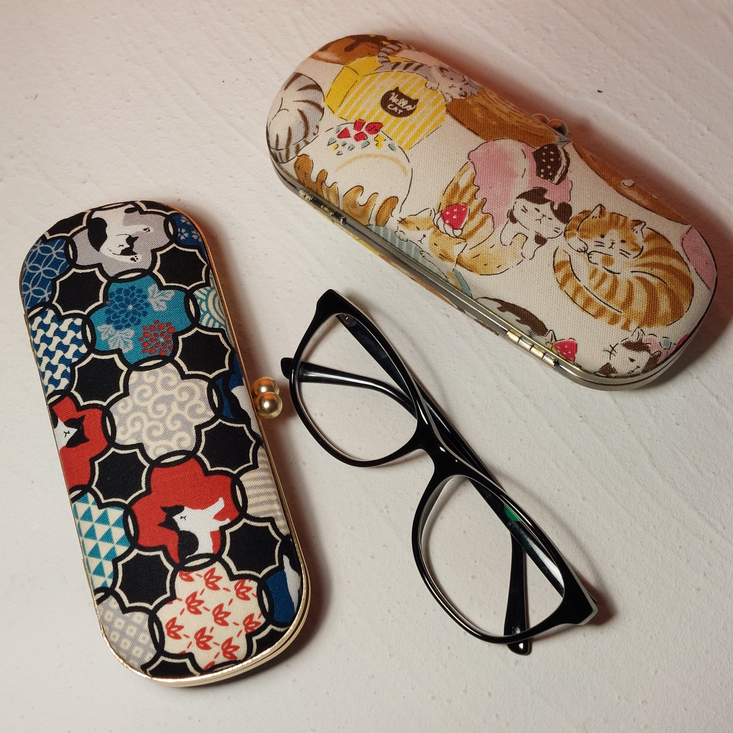 Two handmade fabric hard-shell glasses cases with a pair of black eyeglasses. Left case features a traditional Japanese floral geometric print in navy, red, and cream with a gold kiss-lock clasp. Right case features a whimsical cat and dessert theme.