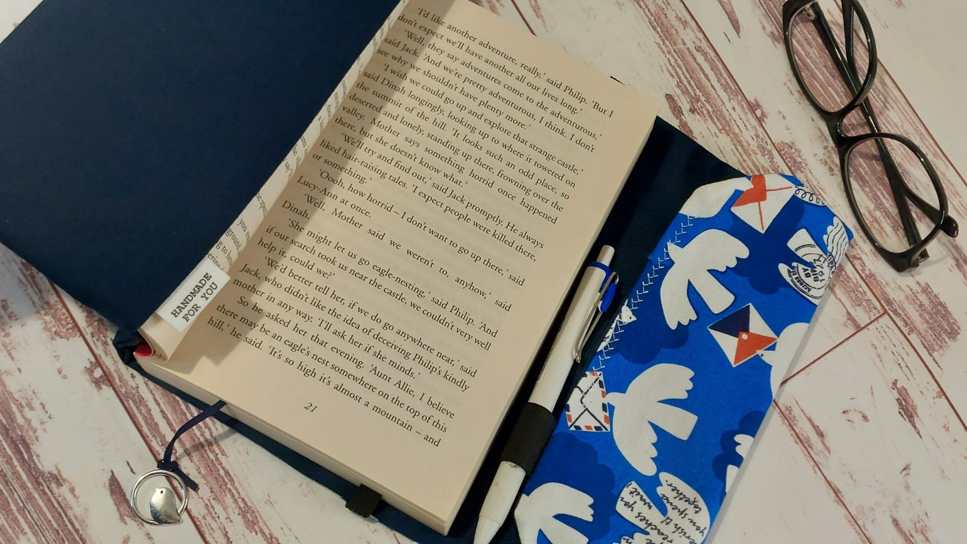 Handmade fabric book sleeve in navy blue with a blue, white, and red pattern featuring doves and envelopes, with a "Handmade for You" tag and a small metal bird bookmark charm.