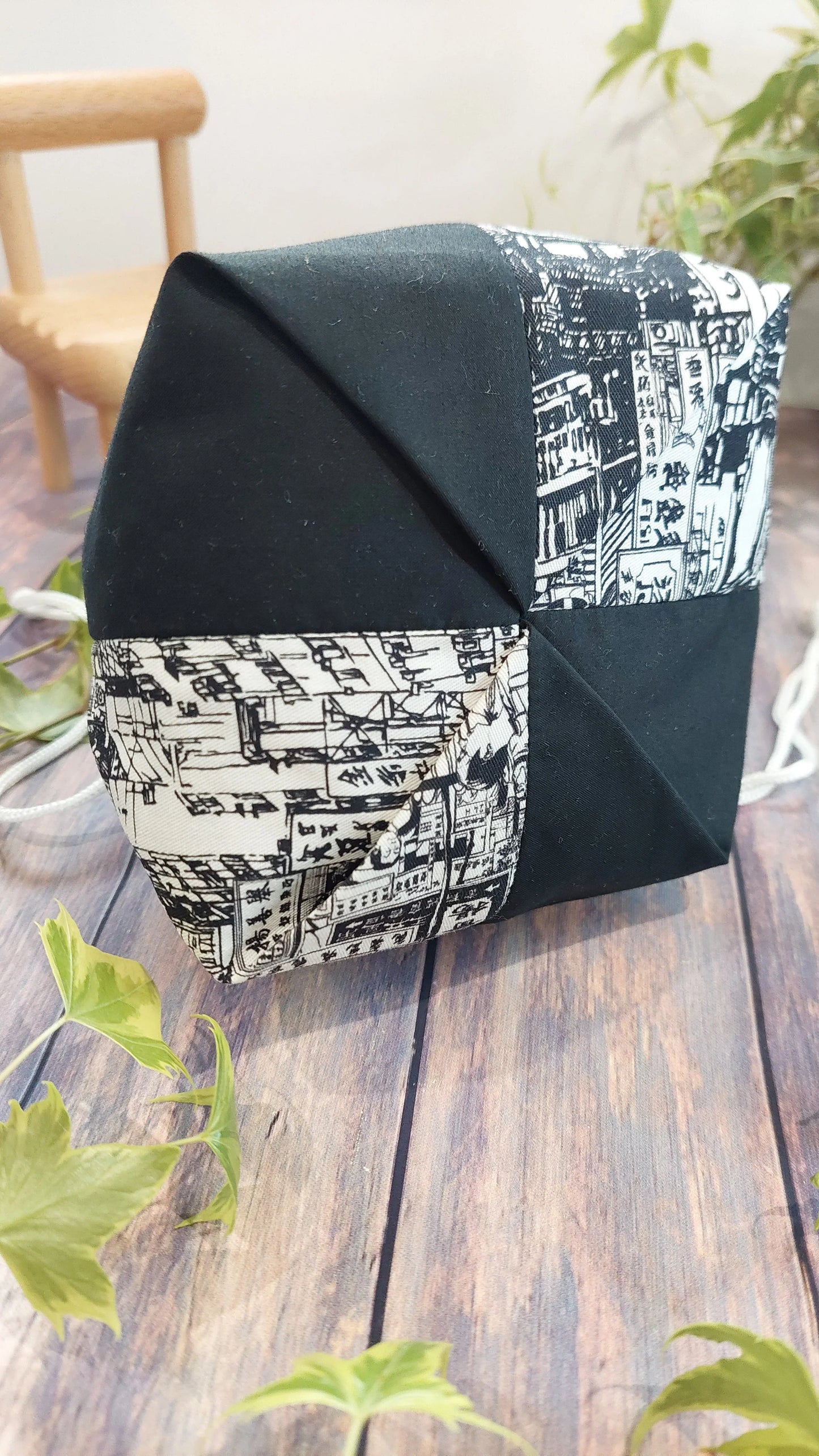 The 折紙風格、拼布的手工抽繩包 displays black and white geometric patterns with intricate line drawings, highlighting 拼接工藝, and is photographed on a wooden surface near green plants.