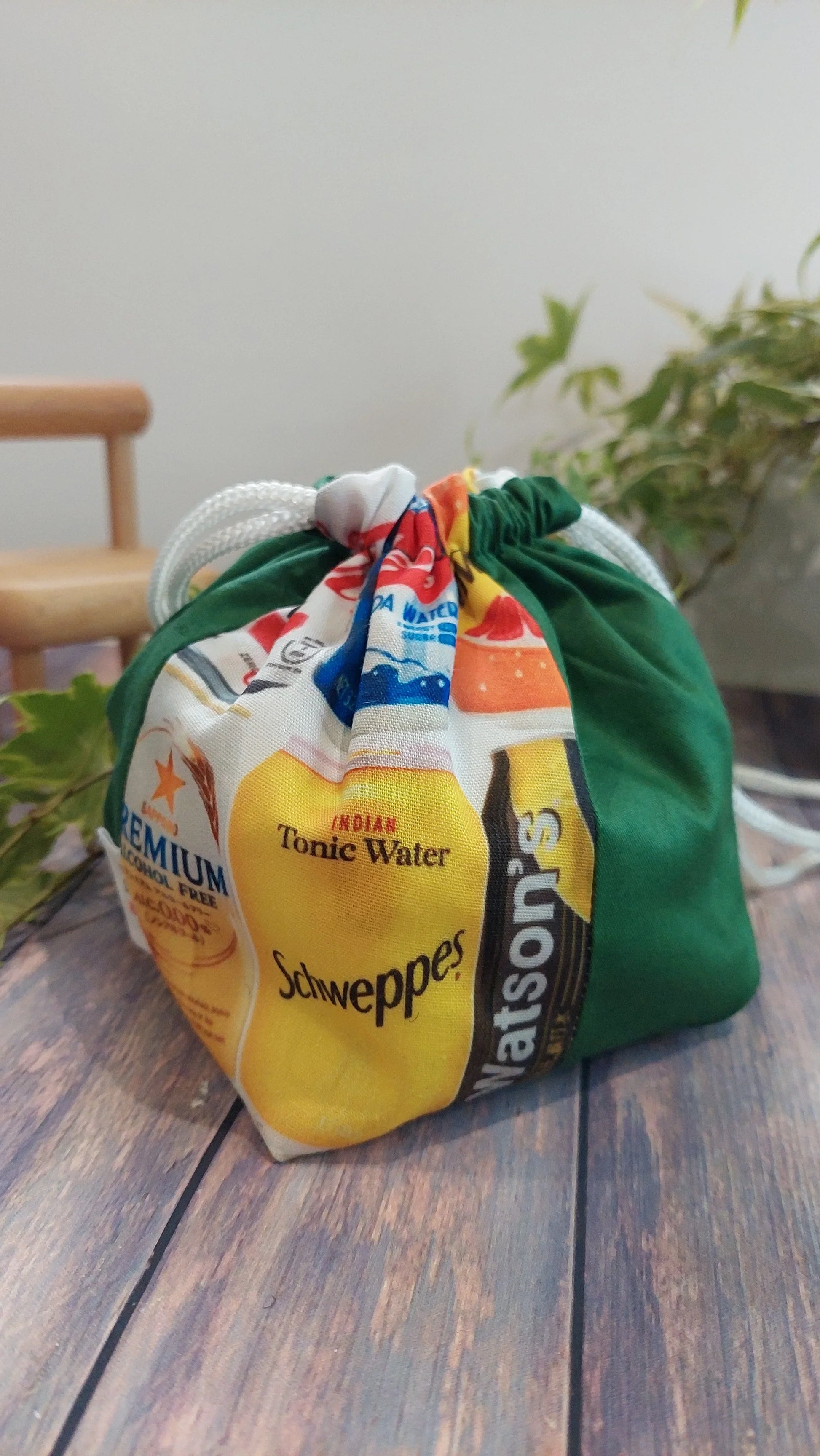 折紙風格、拼布的手工抽繩包以飲品標籤和湯力水印花裝飾，融合拼接工藝，色彩繽紛地陳列在木質表面，背景綠植陪襯。.